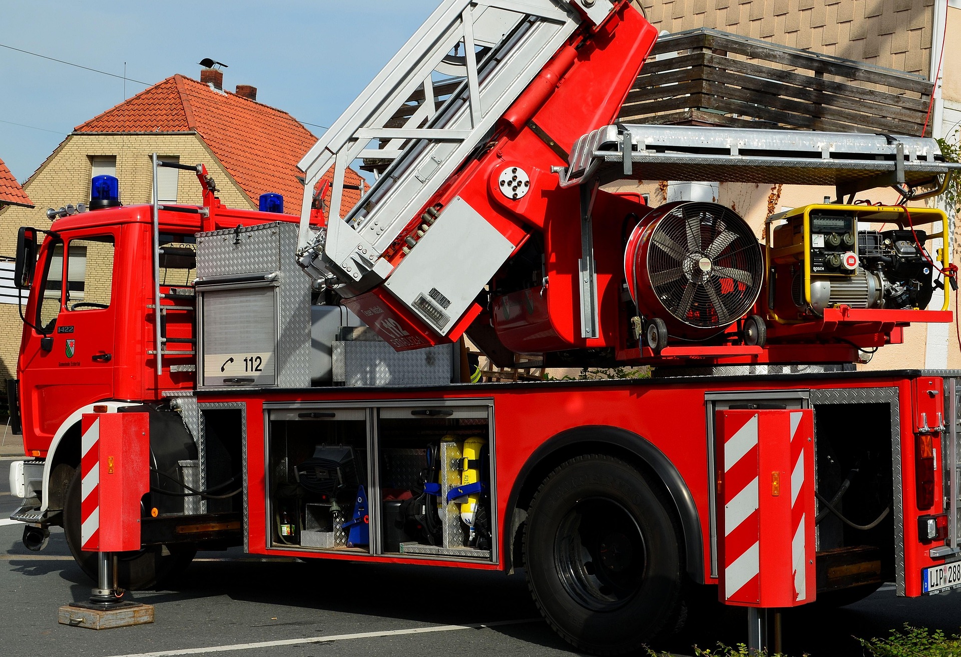 Red aerial ladder fire truck with its stabilizing outriggers deployed and extendable ladder mounted on top, parked on a residential street during an emergency response demonstration.