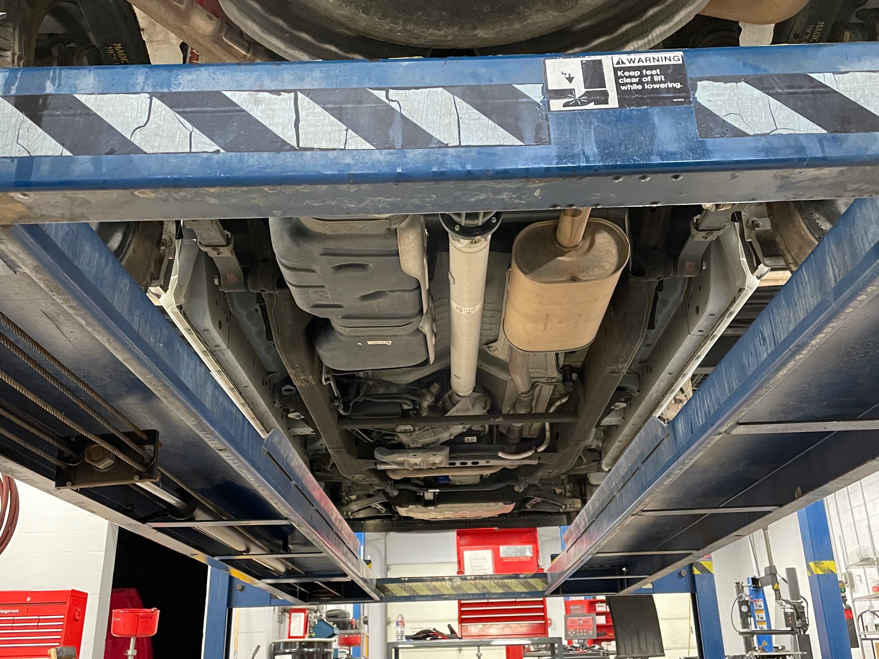 Underside view of a pickup truck on a vehicle lift, showing the factory exhaust system, muffler, driveshaft, fuel tank, and chassis components during an exhaust inspection or installation.
