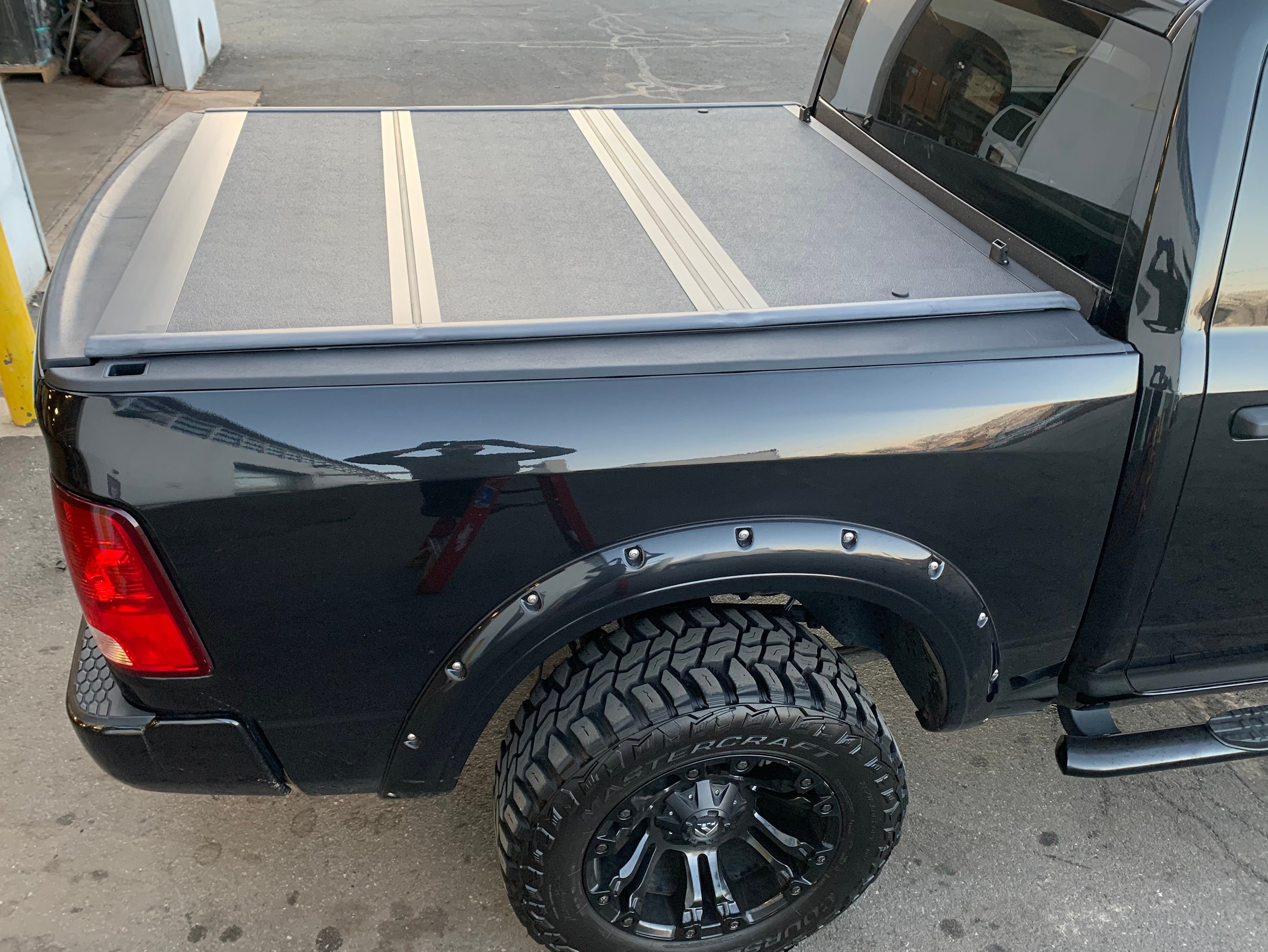 Black Ram pickup with a hard tri-fold tonneau cover closed over the truck bed, featuring rigid aluminum panels and a flush, low-profile design.