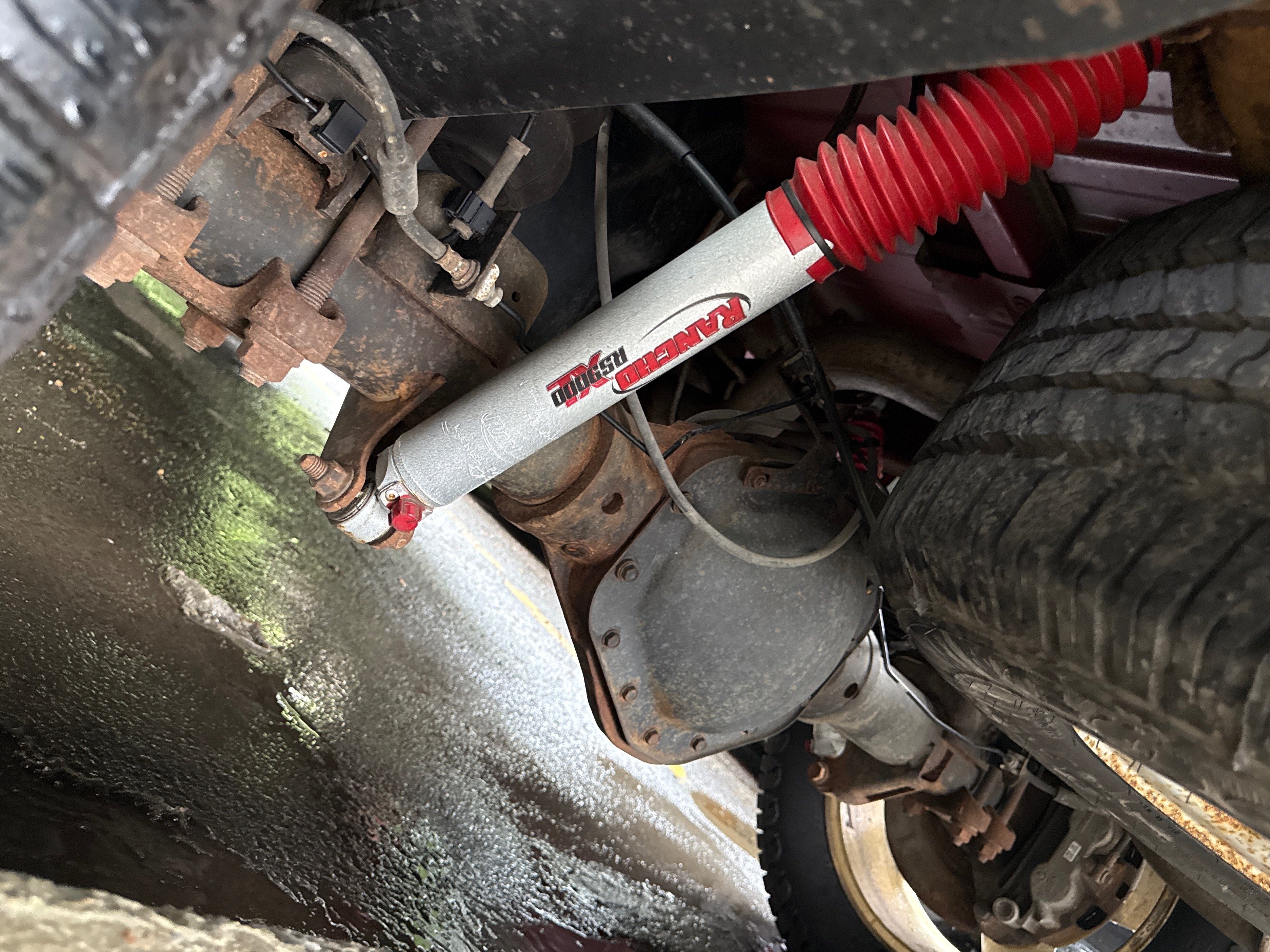 Rancho RS9000XL adjustable rear shock absorber installed on a pickup truck's rear suspension, mounted between the axle and frame to improve ride control, towing stability, and off-road performance.