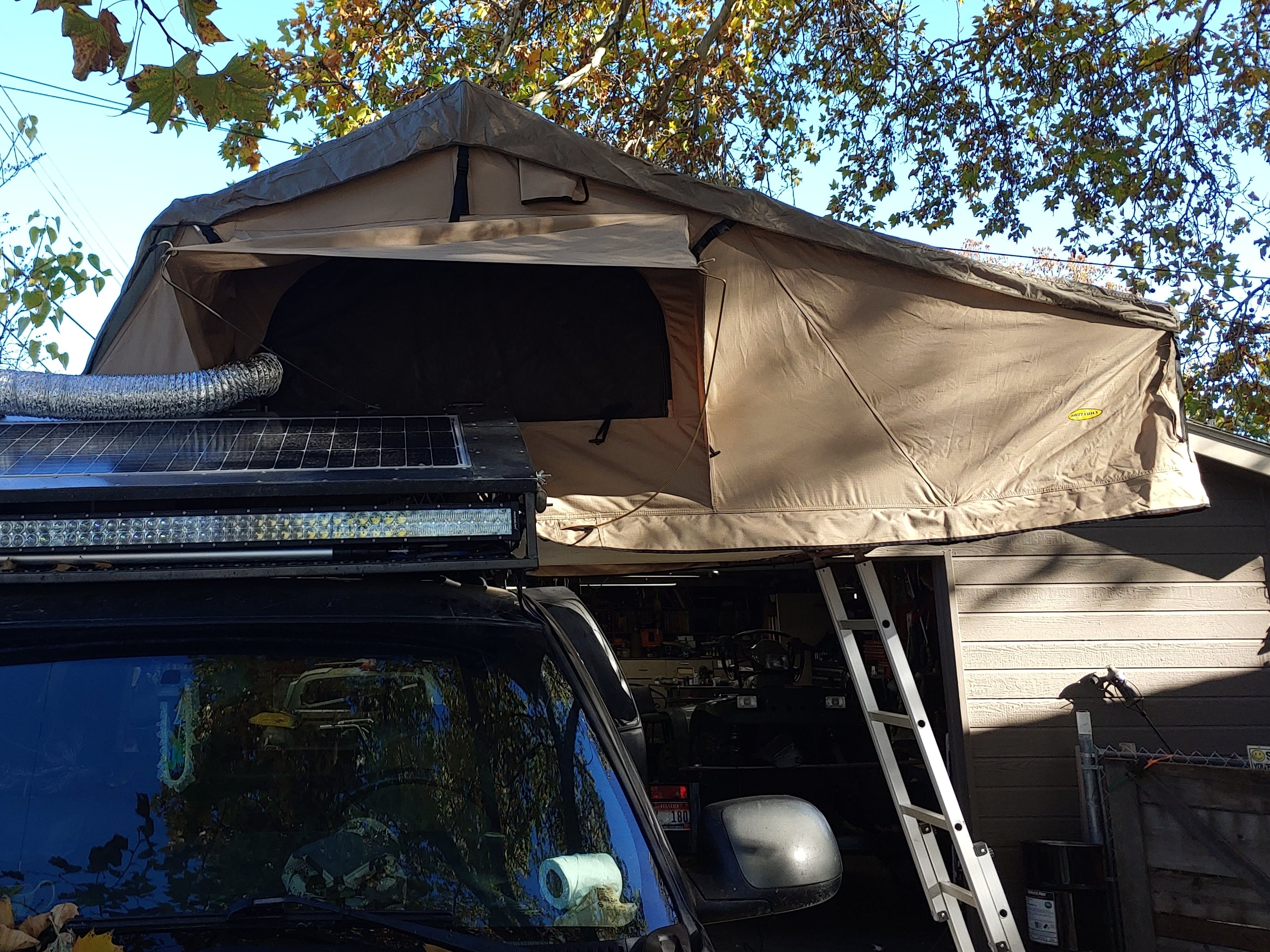Roof-top tent mounted on an off-road SUV with solar panels and ladder setup, parked beside a garage under autumn trees.