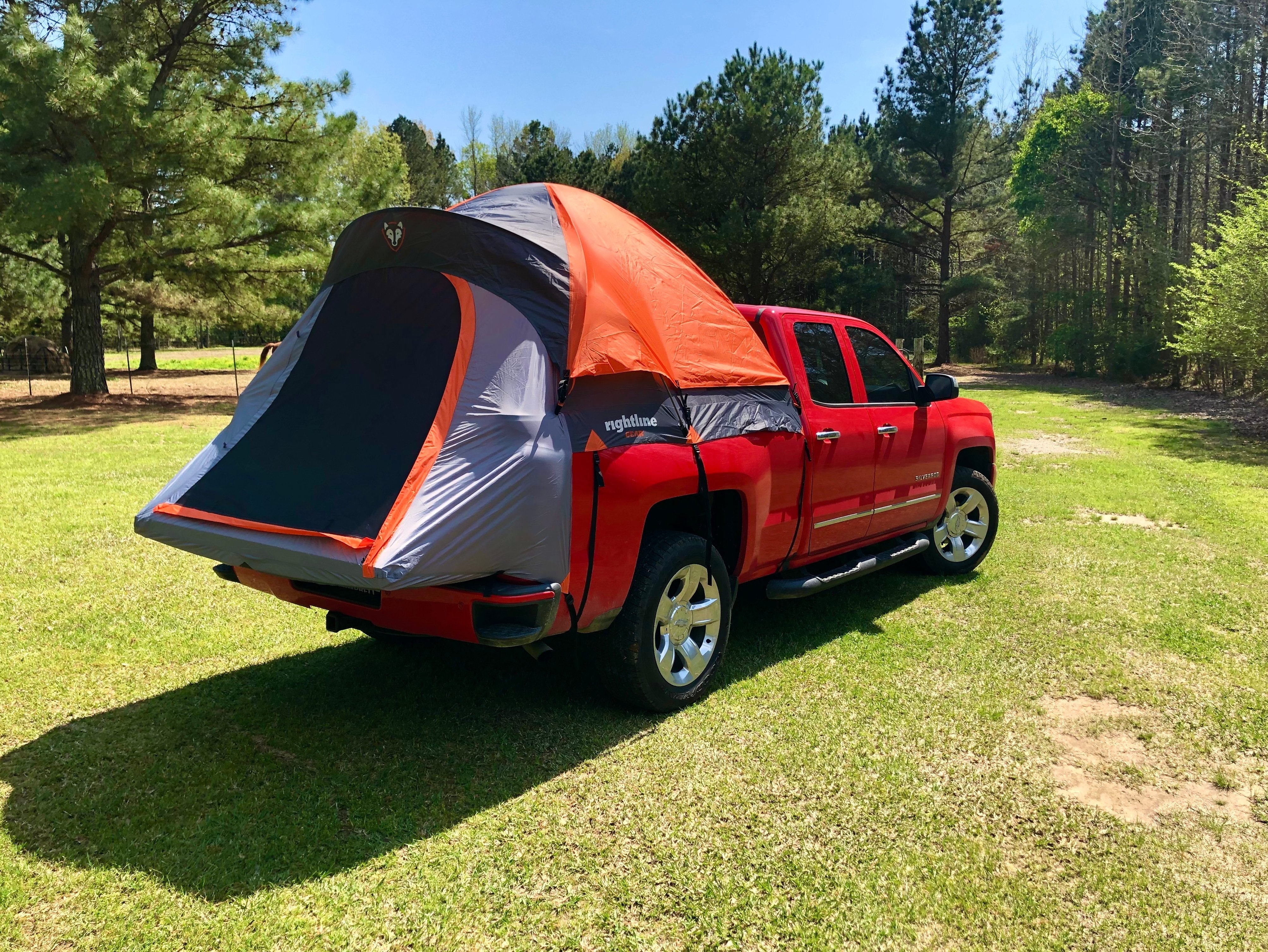 Red pickup truck with a truck bed tent setup parked on a grassy campsite surrounded by trees on a sunny day.