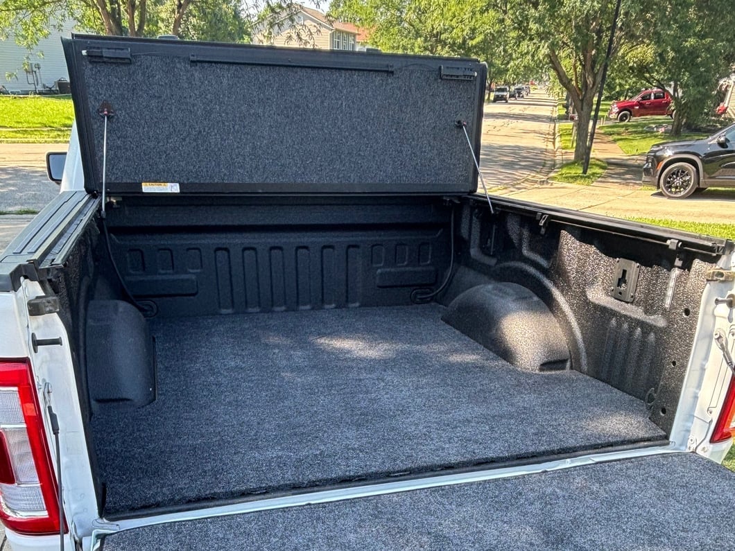 Pickup truck bed with a carpet bed liner installed, featuring a soft, padded surface and a raised one-piece hinged tonneau cover for protected cargo storage.