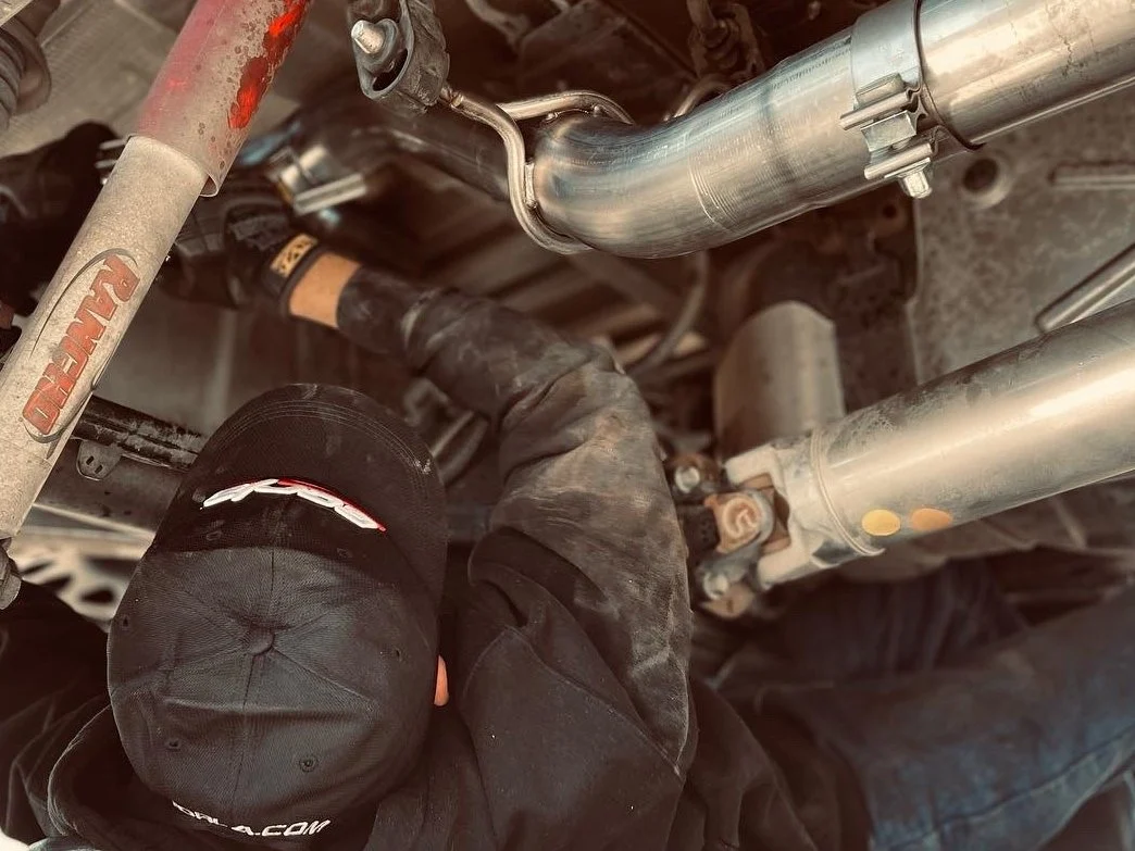 Technician installing a performance exhaust system beneath a pickup truck, tightening stainless steel exhaust tubing and clamps during a cat-back exhaust installation.
